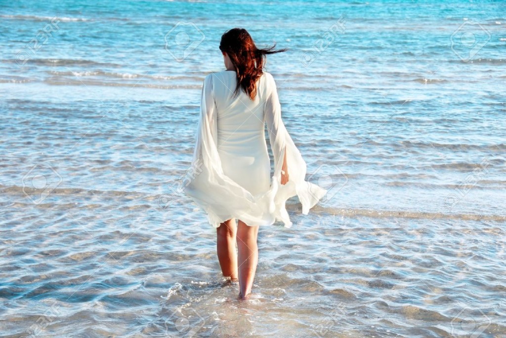 7595990-woman-in-white-dress-and-raised-hands-walking-in-blue-sea-water-Stock-Photo