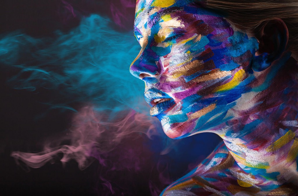 Young woman with colorful make-up and body art on a black background with multi-colored smoke